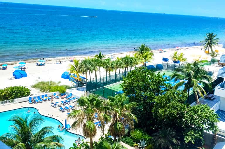 Beachfront resort with blue lounge chairs, pool, sports facilities, palm trees, and ocean view