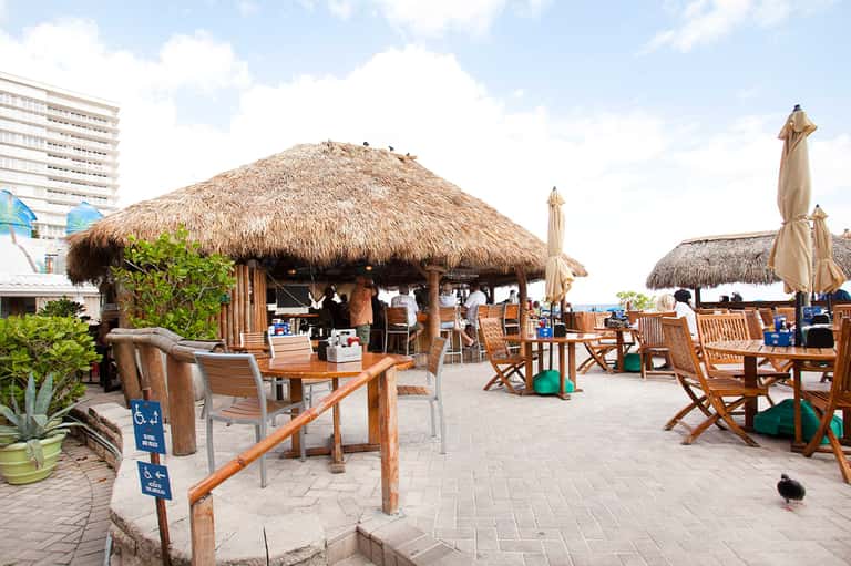 Beachfront tiki bar with thatched roof, wooden furniture, and casual dining area near hotel building