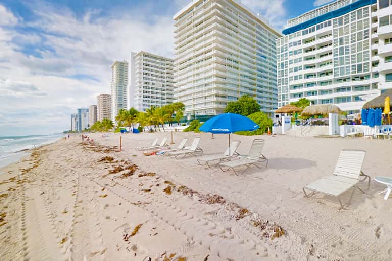 Beach scene with loungers and blue umbrellas, high-rise hotels, palm trees, and ocean