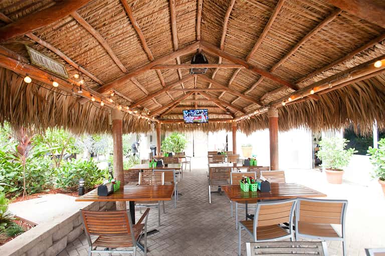 Covered outdoor dining pavilion with thatched roof, wooden tables, chairs, and tropical landscaping