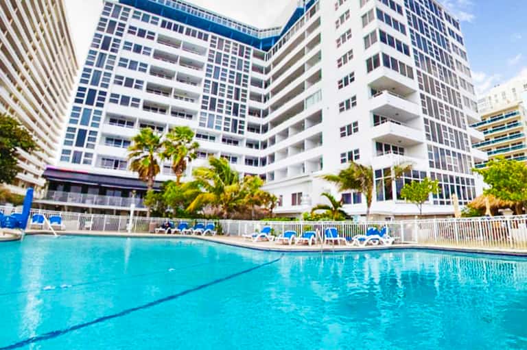 Resort swimming pool surrounded by high-rise residential buildings with palm trees and lounge chairs