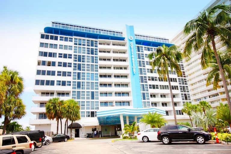 Modern hotel building exterior with blue accents, palm trees, and entrance driveway