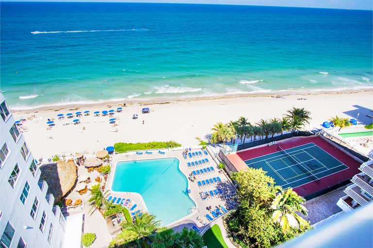 Aerial view of beachfront resort with turquoise pool, sandy beach, tennis court, and high-rise buildings