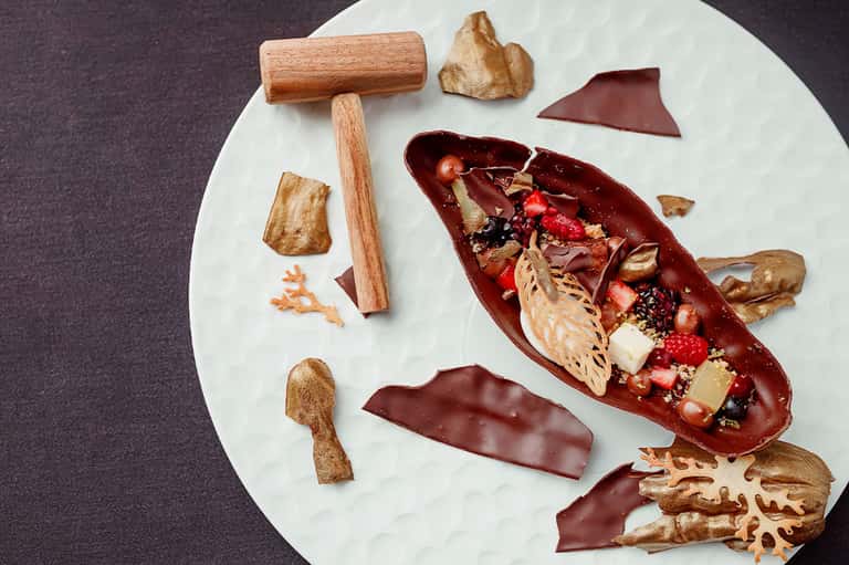 Artistic plated dessert with chocolate elements, fresh strawberries, gold leaf, and decorative wooden pieces