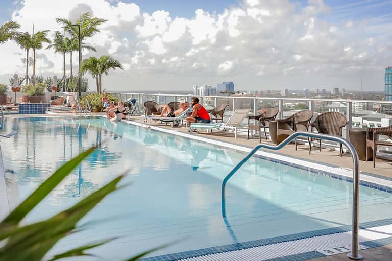 Modern rooftop pool with city skyline views, lounge chairs, and palm trees on sunny day