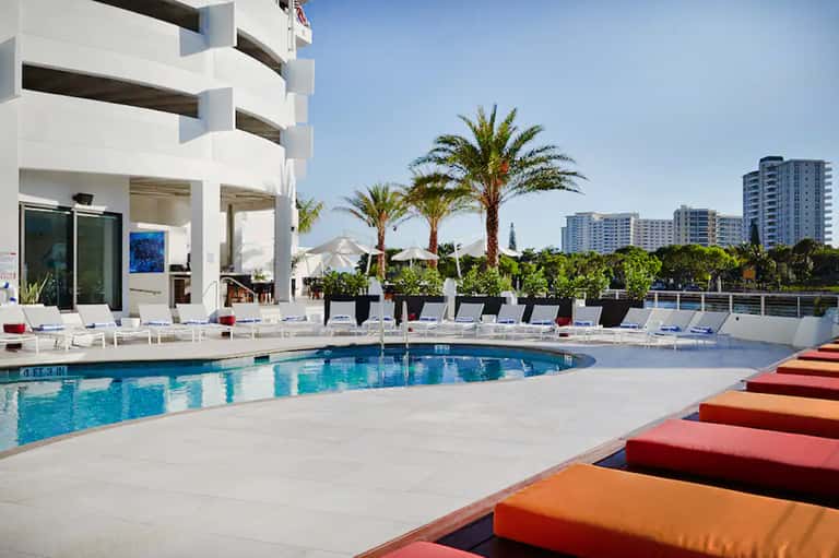 Contemporary pool deck with colorful lounge cushions, palm trees, white building, and highrise hotels