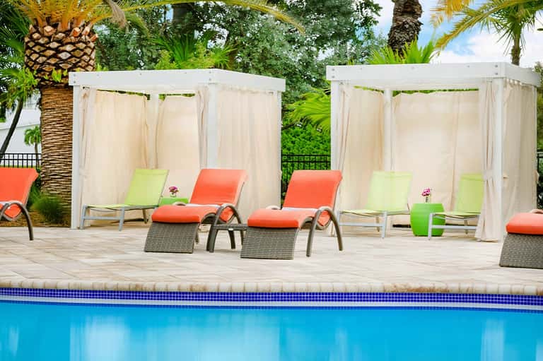 Resort pool deck with orange and lime lounge chairs under white cabanas surrounded by palms