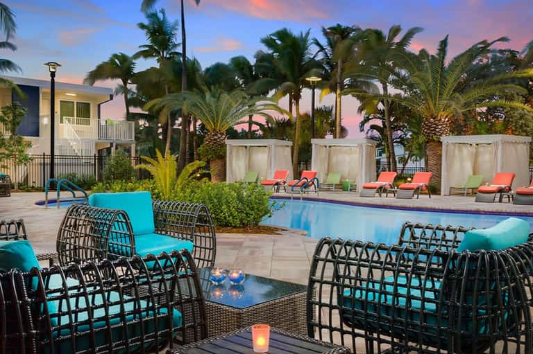 Resort pool deck at dusk with turquoise chairs, palm trees, and illuminated building in background