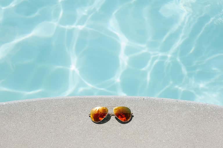 Pair of rose-gold aviator sunglasses on sand with turquoise pool water background