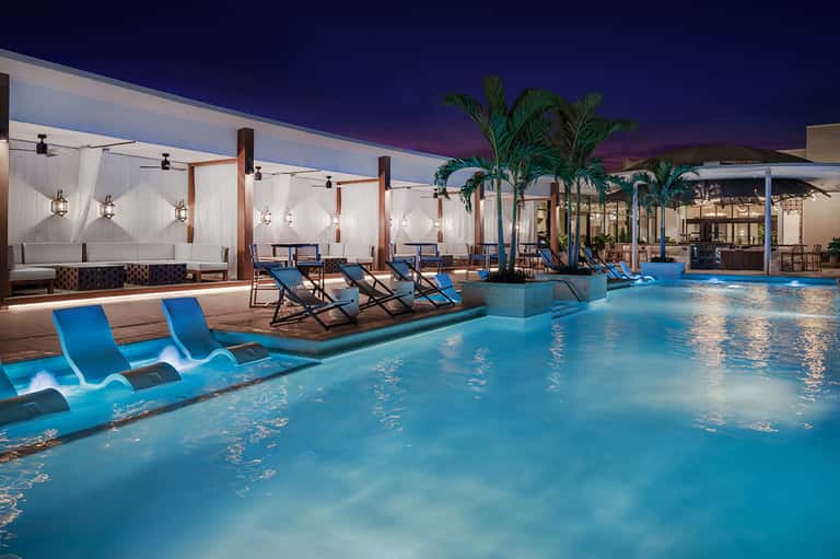 Resort pool at dusk with blue loungers, palm trees, and illuminated cabanas