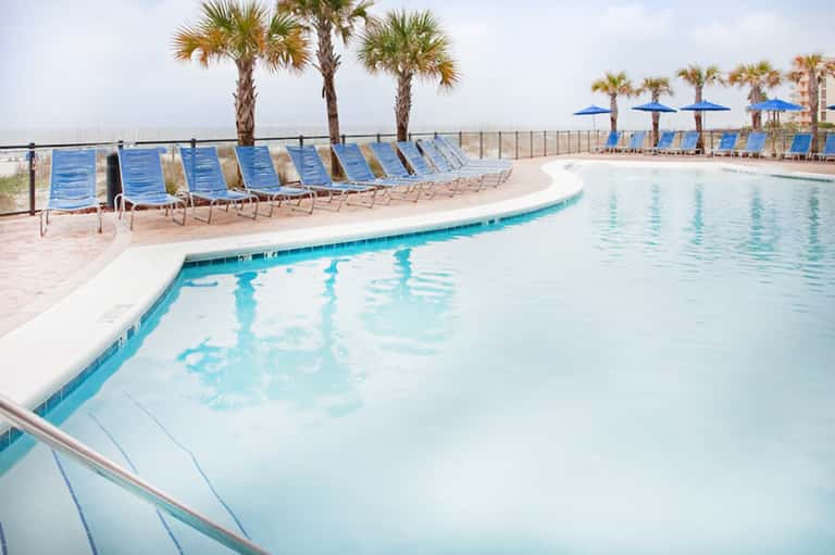 Beachfront pool with blue lounge chairs, palm trees, and ocean view