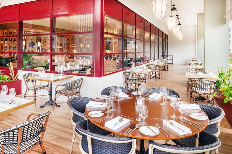 Elegant restaurant with red-framed glass kitchen, wicker chairs, round tables, and wooden flooring