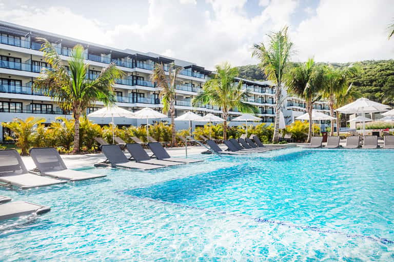 Modern resort pool with turquoise water, lounge chairs, white umbrellas, and palm trees with hillside backdrop