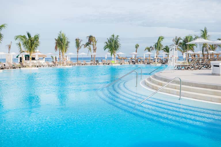 Resort-style pool with palm trees, lounge chairs, and beach in background