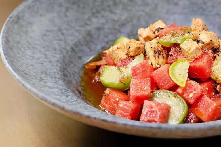 Gourmet appetizer plate with watermelon, croutons, and garnish in gray ceramic bowl