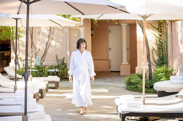 Woman in white robe walking through spa courtyard with lounge chairs and umbrellas