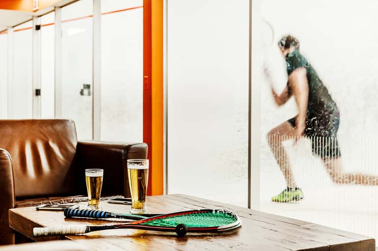 Indoor lounge with leather seating, tennis rackets, beers on wooden table, and squash player visible through glass