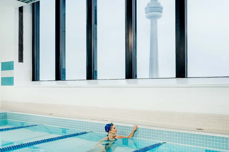 Indoor Olympic-size swimming pool with large windows showing CN Tower Toronto skyline