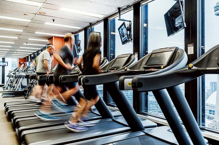 Multiple guests running on treadmills at modern gym with floor-to-ceiling windows and city views.