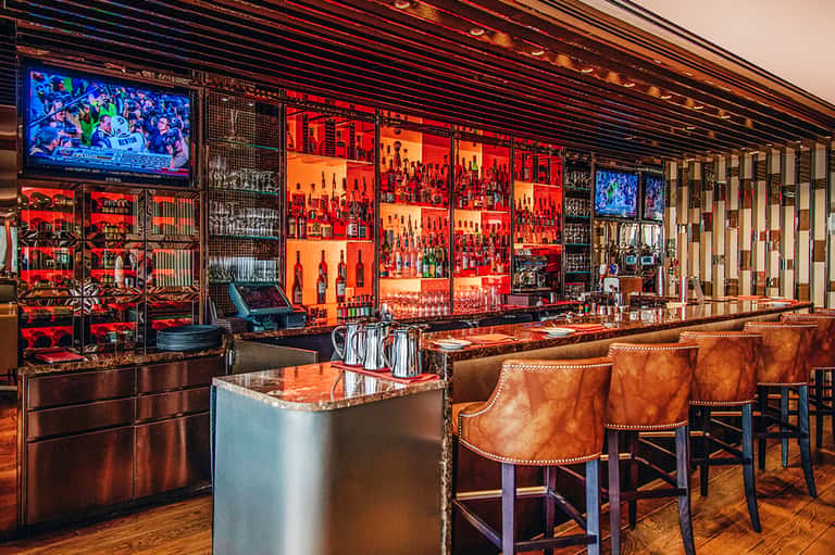 Upscale bar with red and orange illuminated bottle display, leather bar stools, and flat-screen TVs