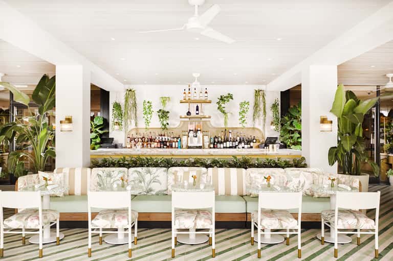 Modern bar lounge with hanging plants, white chairs, green striped bench, and botanical décor