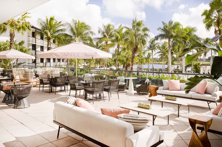 Upscale terrace with contemporary seating, pink cushions, umbrellas, and palm-lined waterfront setting