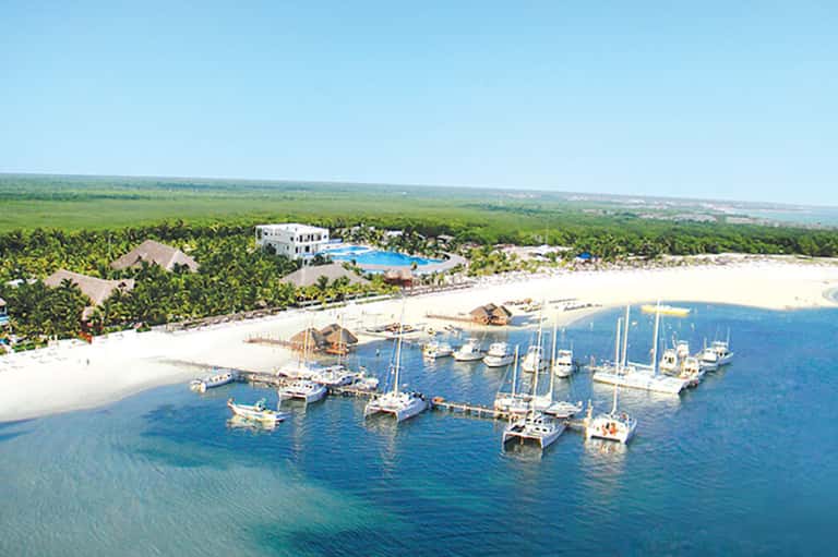 Aerial view of coastal resort with white buildings, pool, boats in marina, and tropical vegetation