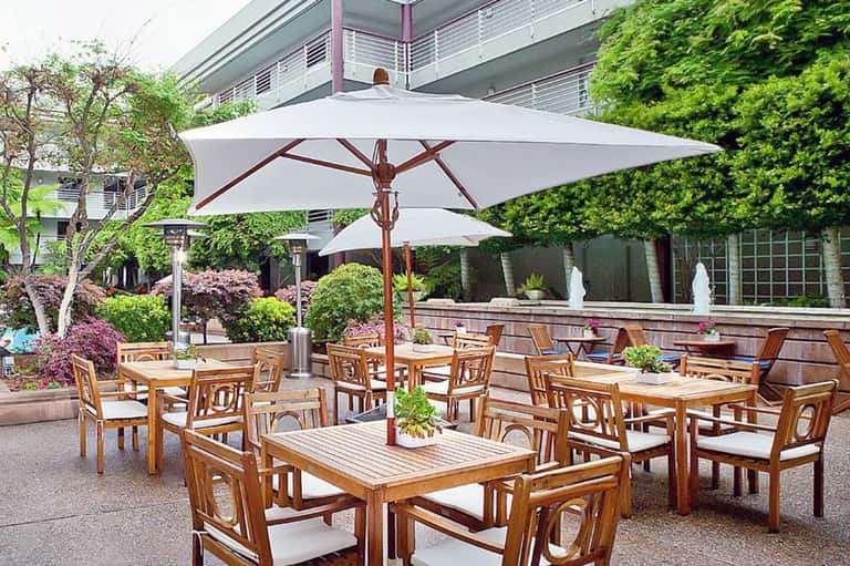 Outdoor patio dining area with wooden tables and chairs under large white umbrellas and green landscaping