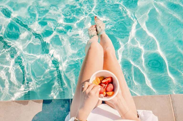 Person's legs dangling above turquoise pool water while holding a bowl of fresh strawberries