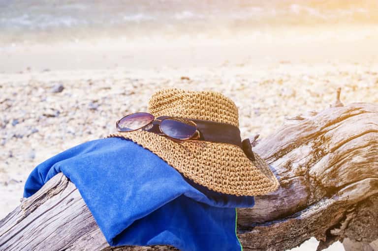 Beach vacation essentials: straw hat with sunglasses and blue towel on sandy shore with driftwood