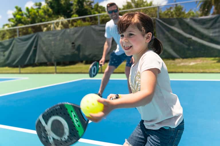Young girl smiling while playing tennis on blue court with instructor in background
