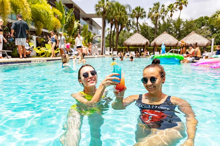 Two guests swimming in bright blue pool with floats, palm trees and thatched umbrellas visible