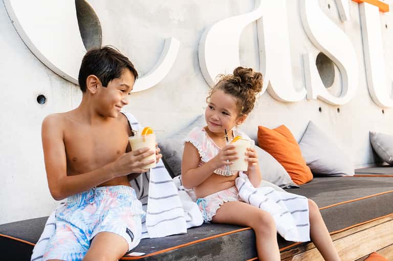 Two children enjoying beverages poolside against white wall with decorative circular design