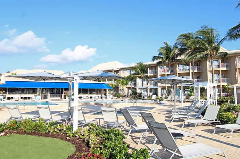 Resort pool deck with loungers under umbrellas, tropical landscaping, and multi-story hotel building