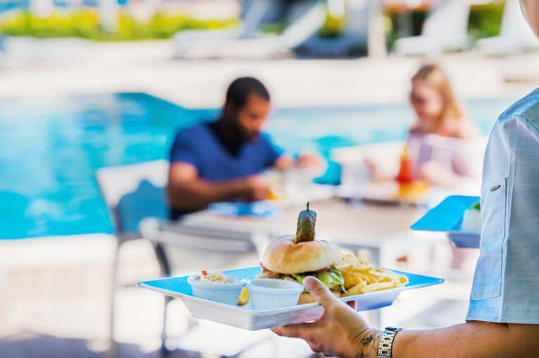 Poolside dining service with server holding plate of burger, fries, and appetizers