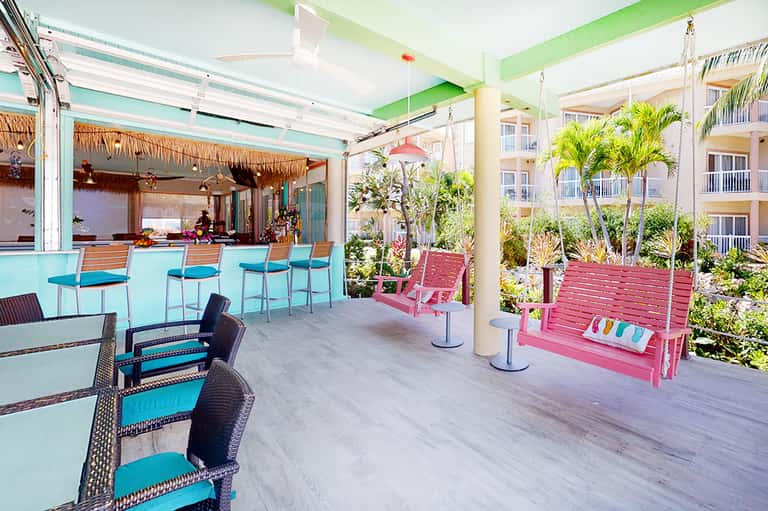Colorful resort bar and lounge with turquoise bar stools, pink swing, potted plants, and tropical decor