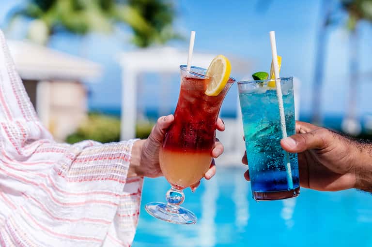 Two colorful cocktails held by hands poolside with ocean view in background