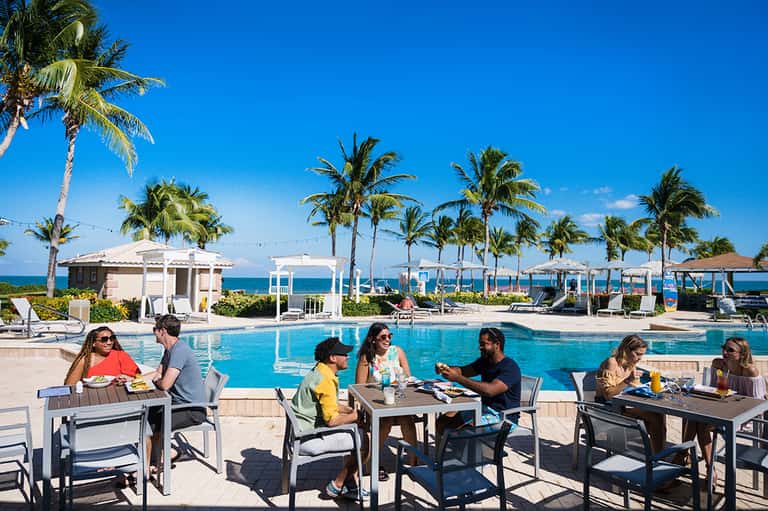 Resort poolside dining area with guests, blue water, cabanas, palm trees, and ocean view