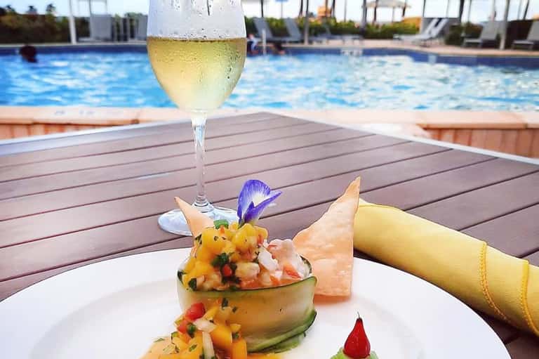 White wine glass and gourmet dish on wooden deck overlooking blue swimming pool and cabanas
