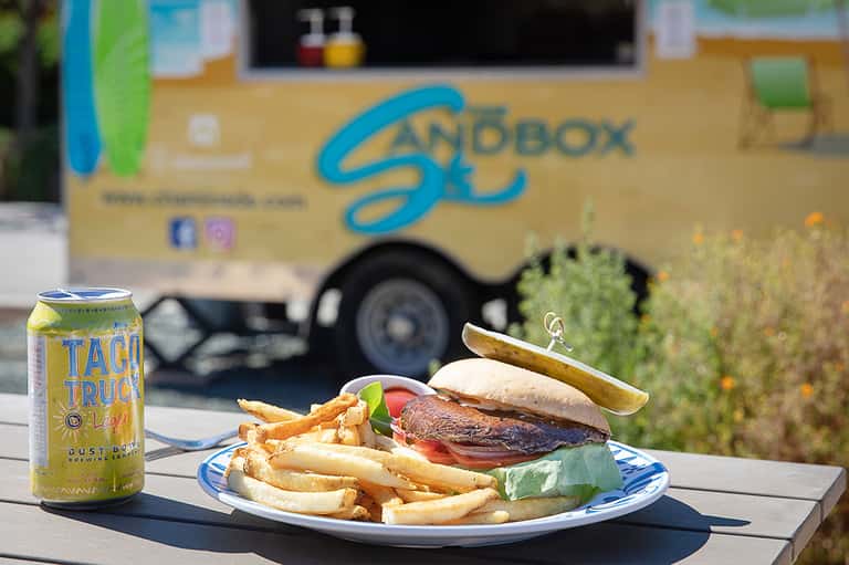 Gourmet burger with fries and beverage in front of Sandbox food truck branding