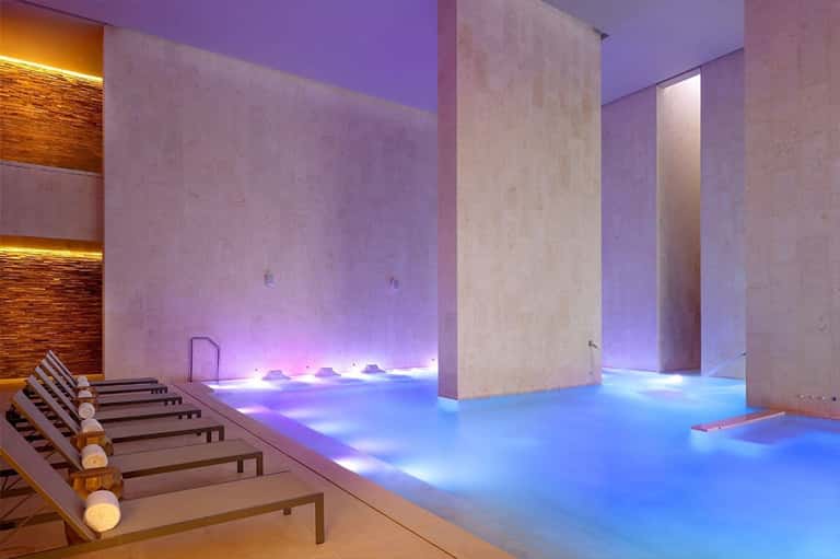 Luxury spa pool with blue lighting, concrete columns, and lounge chairs along the water