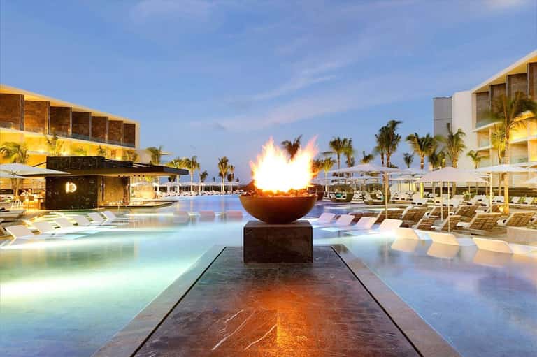 Modern resort pool at dusk with illuminated fire bowl sculpture, loungers, palm trees, and coastal architecture