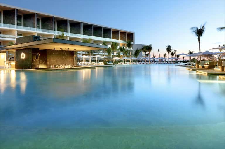 Modern resort pool at dusk with illuminated bar, palm trees, and sleek architecture with glass railings