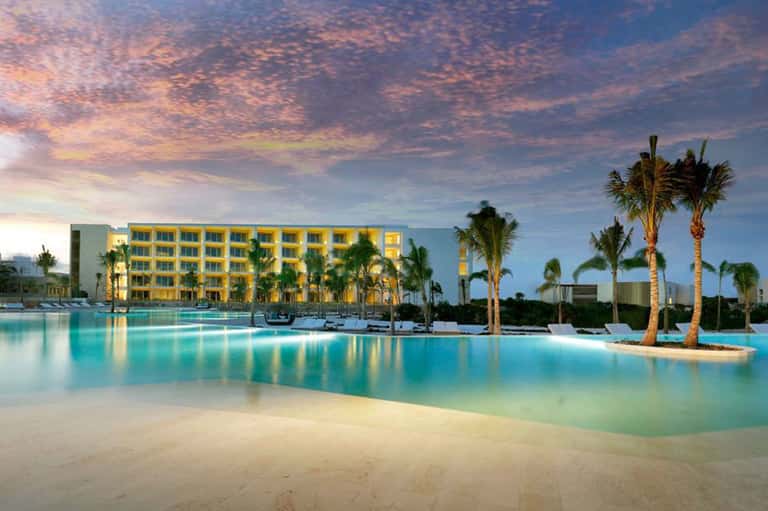 Beachfront resort at sunset with illuminated yellow building, palm trees, and turquoise lagoon pool