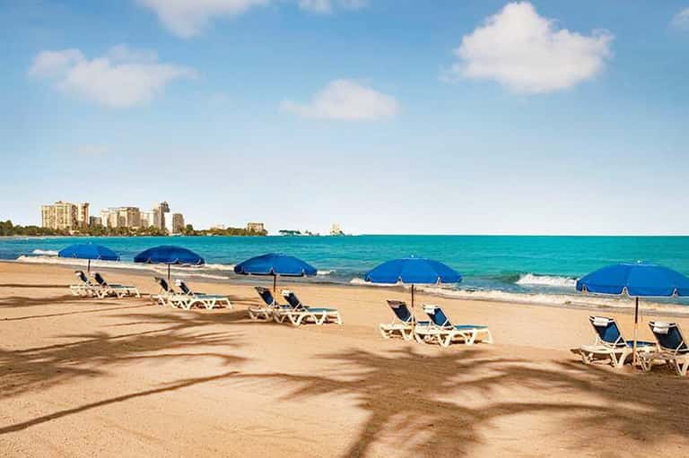 Sandy beach with rows of white lounge chairs, blue umbrellas, and turquoise ocean with coastal buildings in background