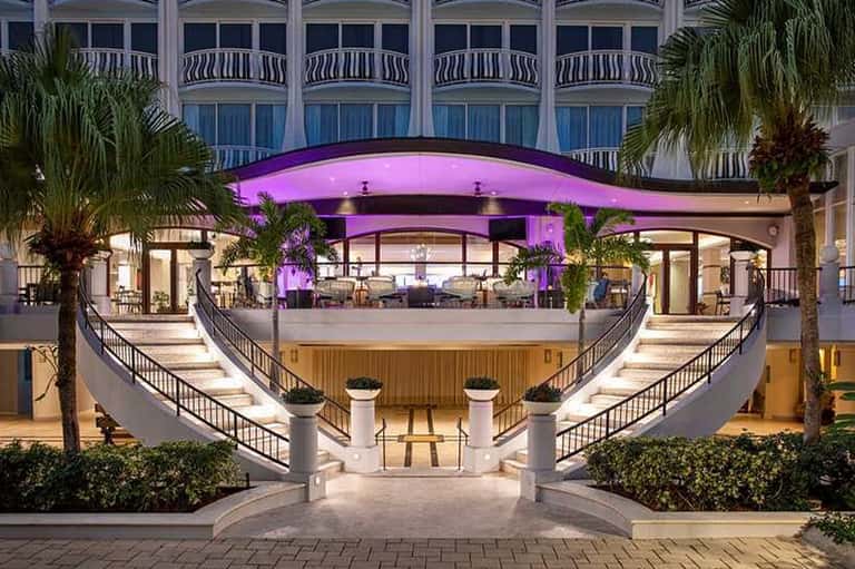 Modern hotel atrium with pink-lit curved bar, symmetrical staircases, and palm tree accents