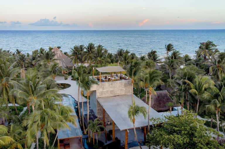 Aerial view of beachfront resort with multi-level structures, pools, and palm trees by ocean