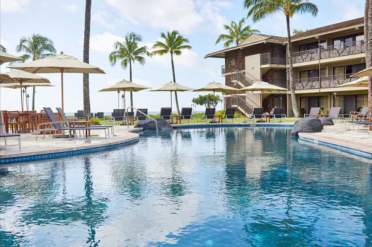 Resort pool with tan umbrellas, lounge seating, palm trees, and ocean view beyond