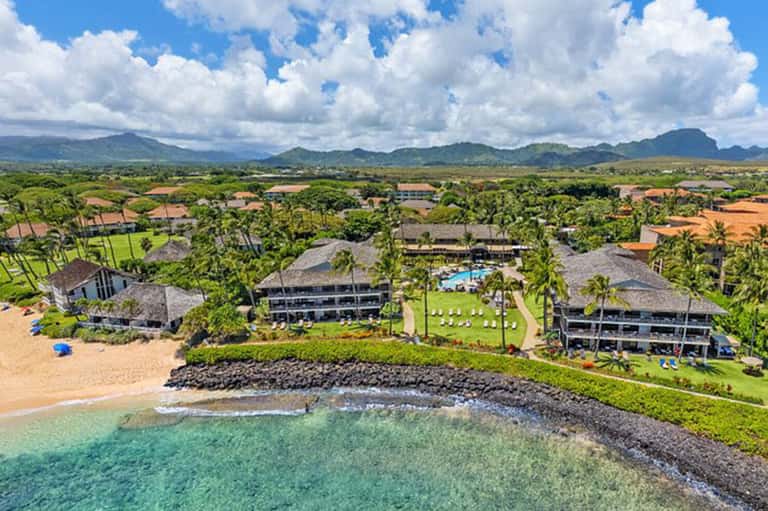 Aerial view of beachfront resort with thatched-roof buildings, pools, and lush grounds on Hawaii coast