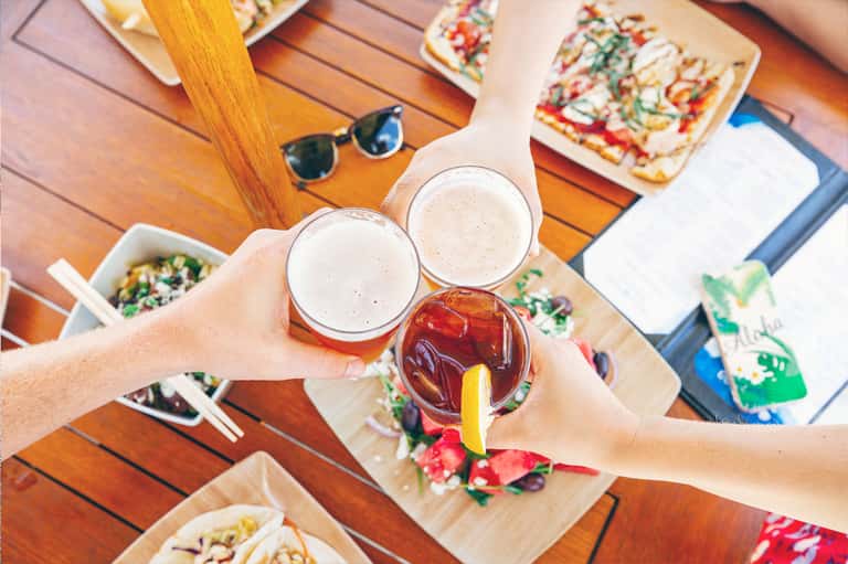 Overhead view of two beer glasses and cocktail with hands toasting at wooden table
